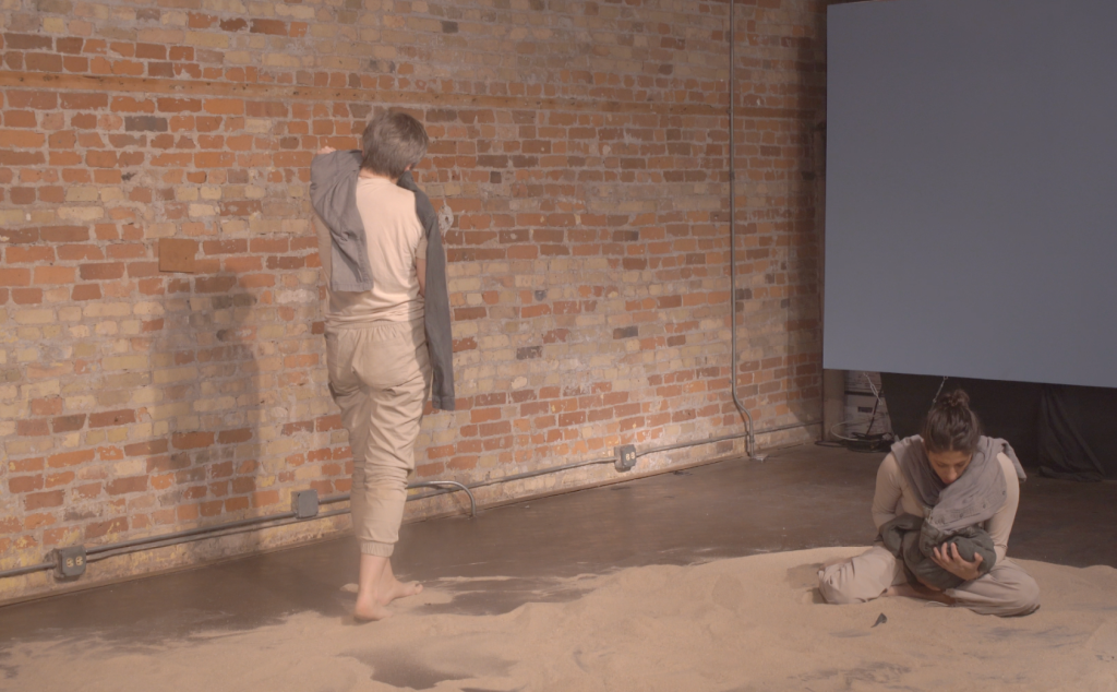 Image: Maria Plotnikova and Sofía Gabriel are shown holding the garments during the John David Mooney Performance of Territory and People. Plotnikova is shown standing towards the back wall while Gabriel is holding the clothes in her hands like as if it were a small child. Image by Mallory Qiu.